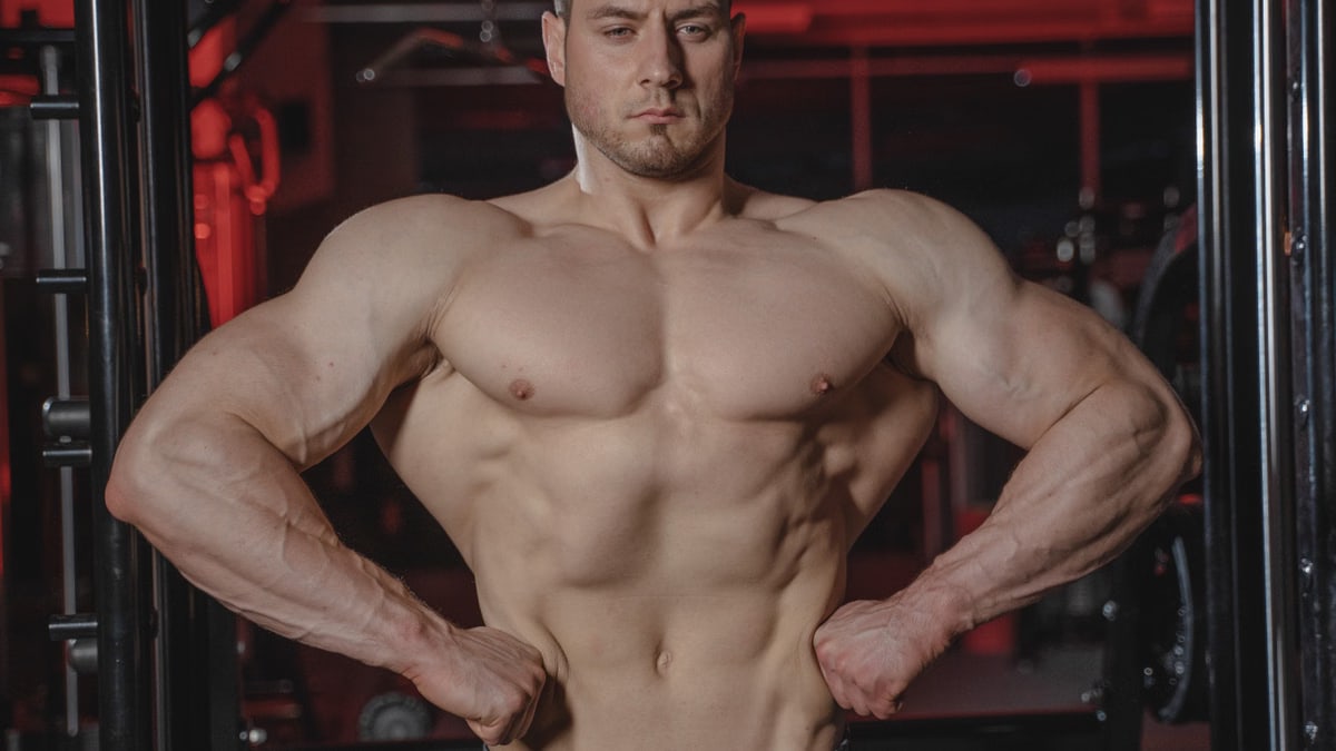 Intense shot of a muscular man posing topless in a gym setting showcasing physique.